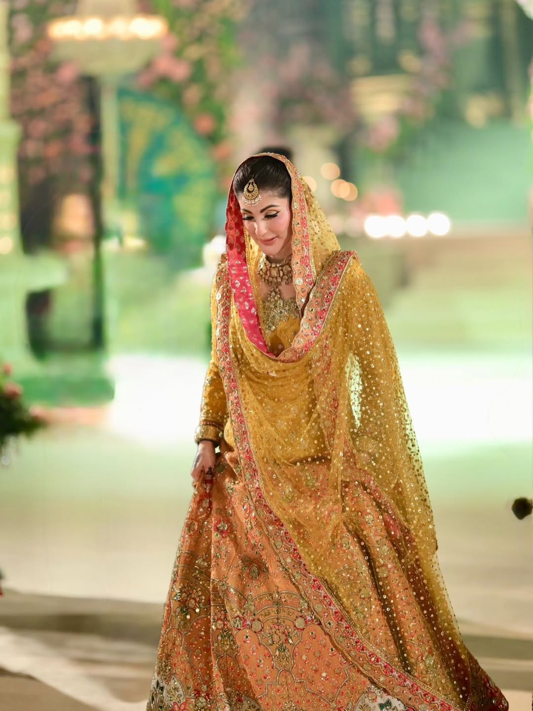 Luxury mustard yellow bridal lehenga choli featuring intricate hand embellishments with detailed floral embroidery in soft pastel hues along the lehenga border, paired with a heavily adorned net dupatta with contrasting pink and gold border and a finely embellished choli, perfect for mehndi, mayun, dholki, and wedding festivities.